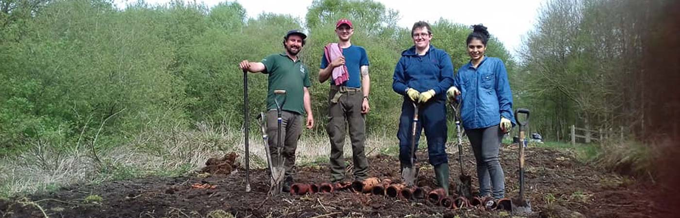 A group of people stand in a muddy field holding spades.