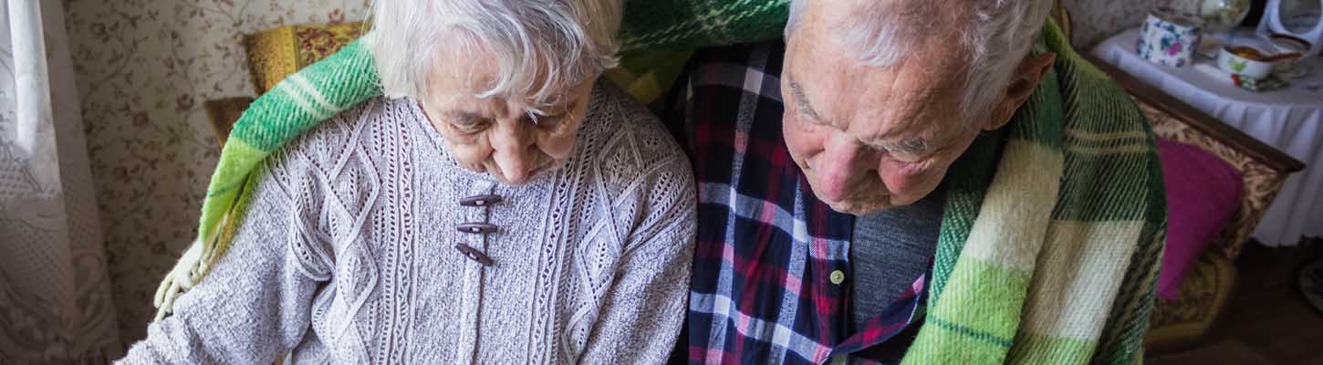 An elderly couple sit in a chair with a knitted blanket around their shoulders, suggesting a cold house.